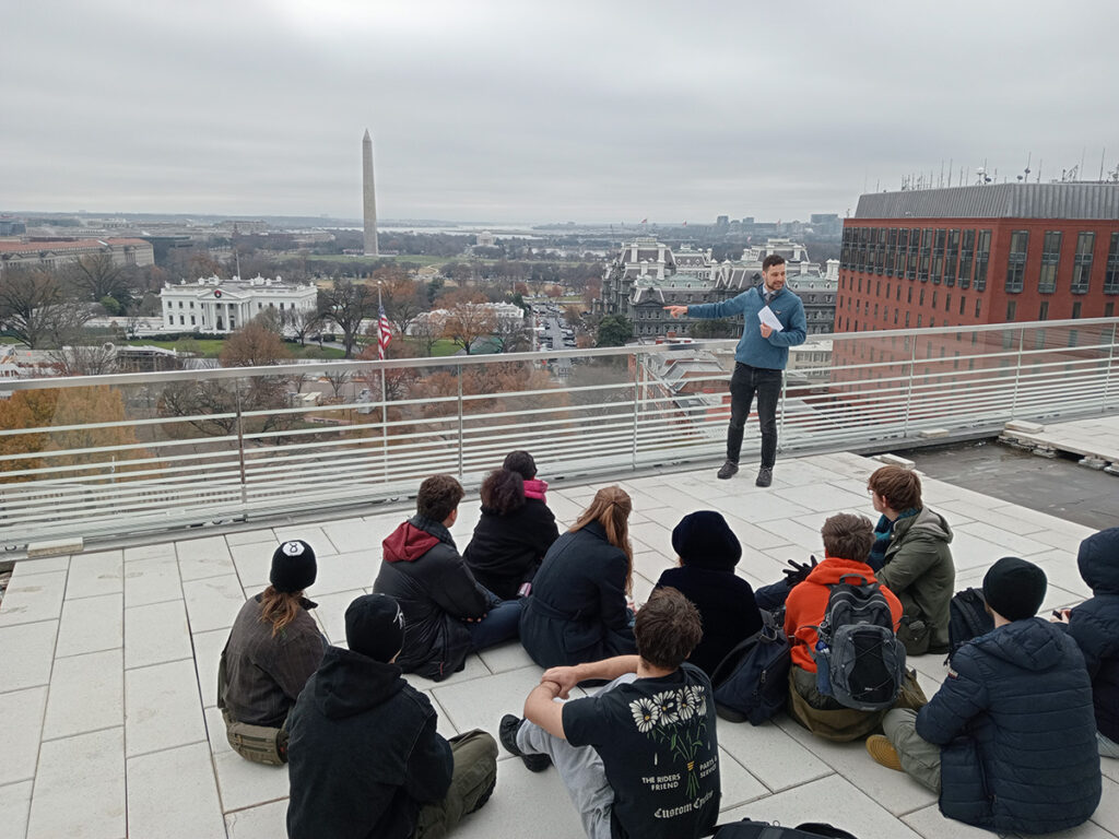 Students sit on a rooftop in Washington DC, with the Washington Monument visible in the background, listening to a male journalist talk