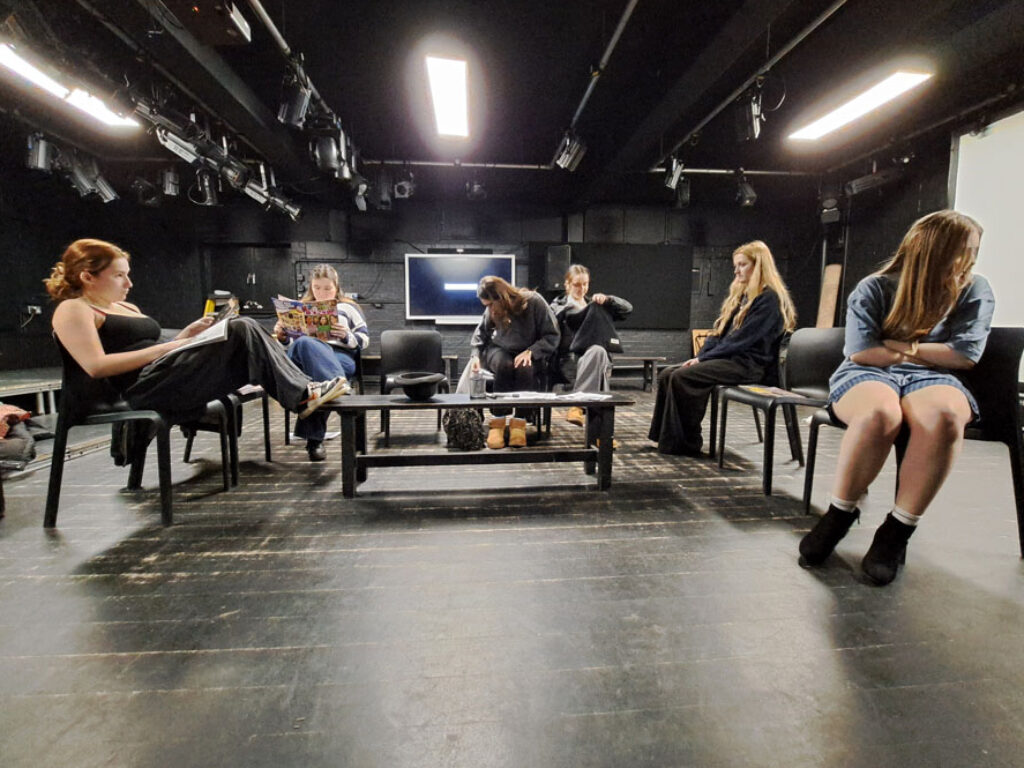 Six students from The King Alfred School North London sit on chairs around a low table as they perform a play in the rehearsal studio