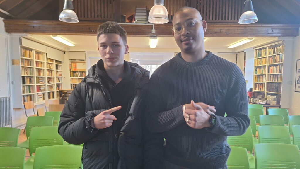 A young white male stands next to a black man wearing glasses in an Old Library