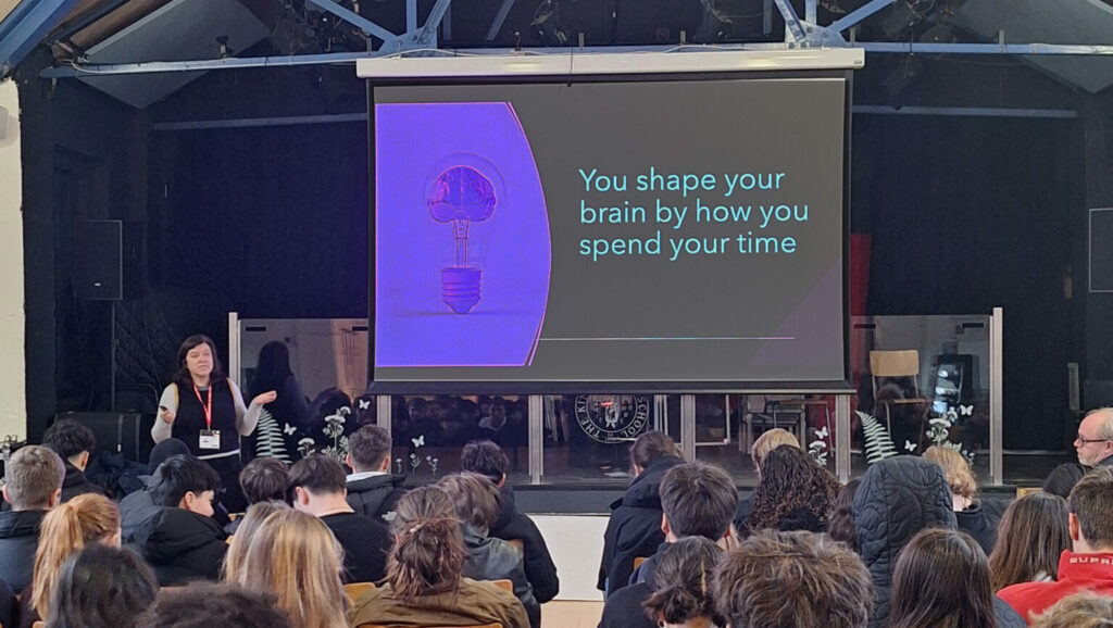 A white woman presents to a room of students in front of a screen showing a slide that reads 'You shape your brain by how you spend your time'