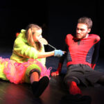 A girl dressed in a tutu and green onesie licks a lolly sat on the floor next to a boy in a spiderman costume as part of a performance on a stage