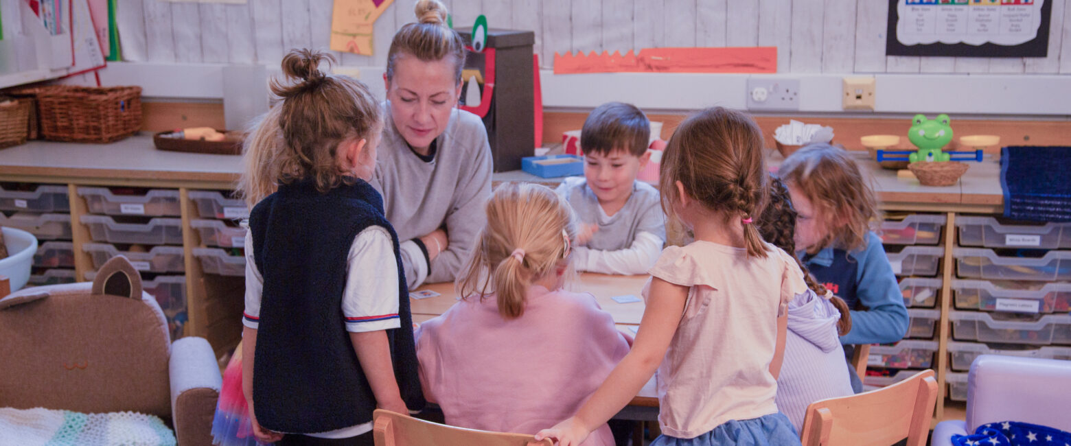 A group of Reception students sit round a table reading with a member of staff at The King Alfred School North London