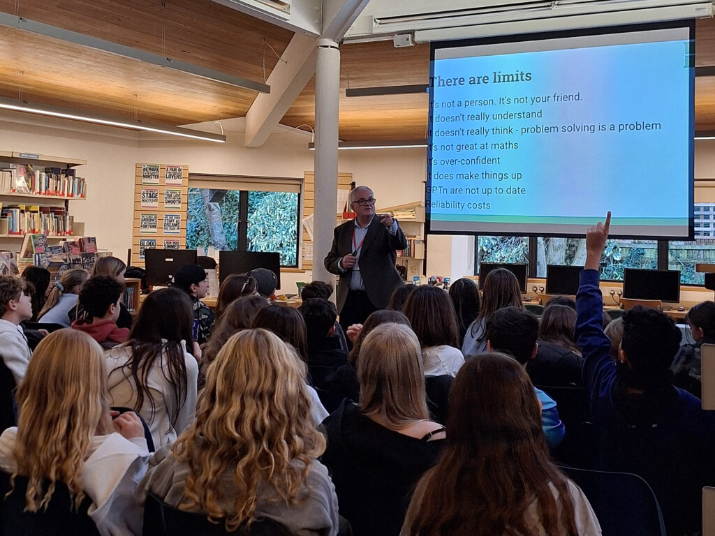 A white male wearing a brown jacket and white shirt stands in front of a room of seated students. He points to a student with their hand raised. Behind him is a screen listing the limitations of ChatGPT for use in study.