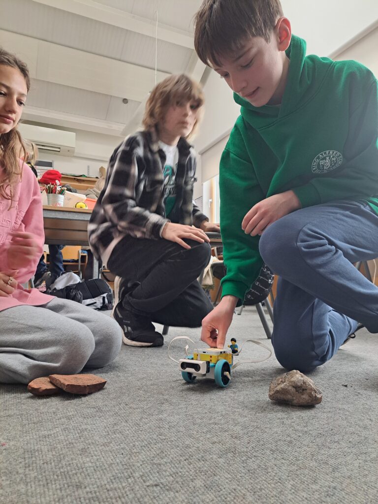 Three students kneel on the floor of a classroom as they test a mini Mars rover made out of Lego, navigating it around small rocks