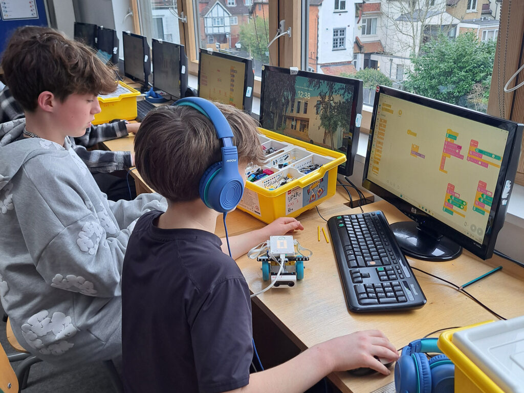 Two students, one wearing blue headphones, sit at a desk, using a computer to use a coding programme