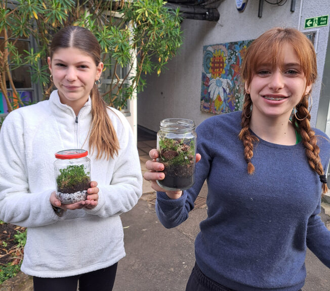 Two female students stand outside holding terrarium in glass jars