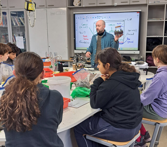 A male teacher holding a terrarium stands in front of students sat at science benches