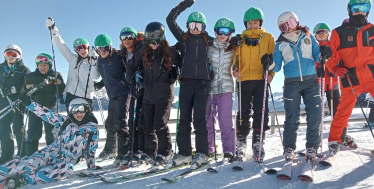 Students wearing skis and snowsuits pose in a line on a snowy mountain